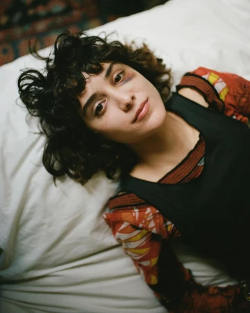 Indoor portrait of a young adult woman with curly brown hair lying on a white bed wearing a black dress over a patterned orange top.