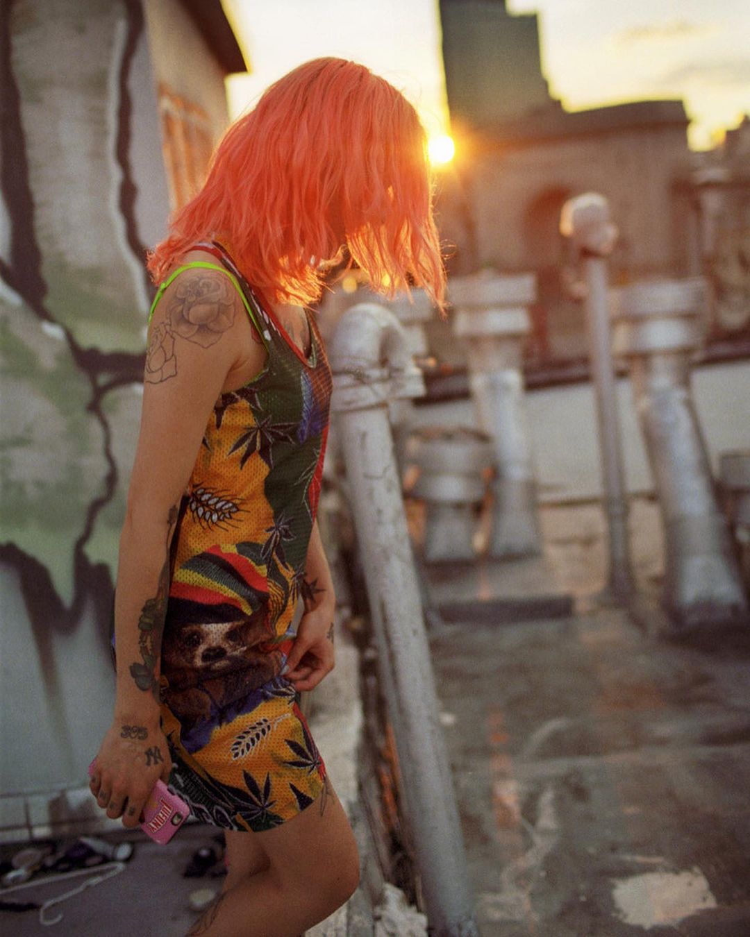 A woman with orange hair stands on a rooftop at sunset, wearing a patterned dress and holding a phone, with city buildings in the background.