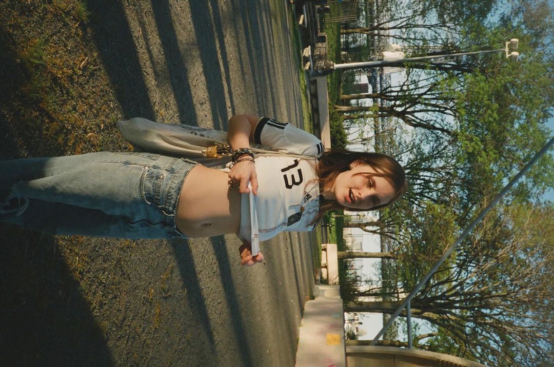 Young adult poses outdoors wearing a sports jersey and jeans, showing her stomach while standing on a paved surface with trees in the background.
