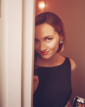 Woman in black top looking around a white door frame with a subtle smile captured in soft warm indoor lighting by Chad Moore.