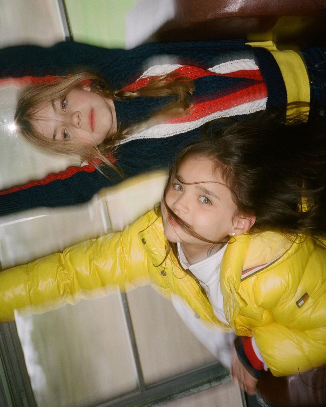 Two young girls lie on their backs inside a vehicle, wearing a yellow puffer jacket and a striped sweater in a cheerful, casual shot.