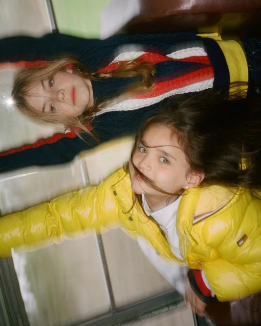 Two young girls lie on their backs inside a vehicle, wearing a yellow puffer jacket and a striped sweater in a cheerful, casual shot.