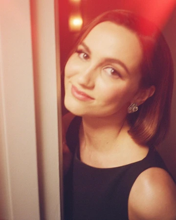 Close-up portrait of smiling woman with short brunette hair and earrings, wearing black sleeveless top, looking at the camera indoors.