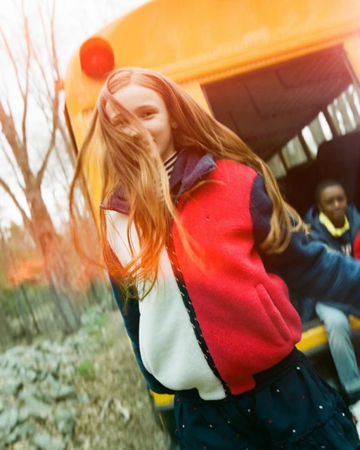 Young girl exits yellow school bus in a red, white, and blue fleece jacket and a patterned skirt, another child is visible in background.