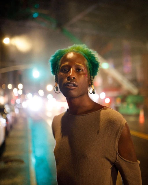 Adult with green hair wearing a tan top and a nose ring stands on a city street at night with blurred cars and lights.