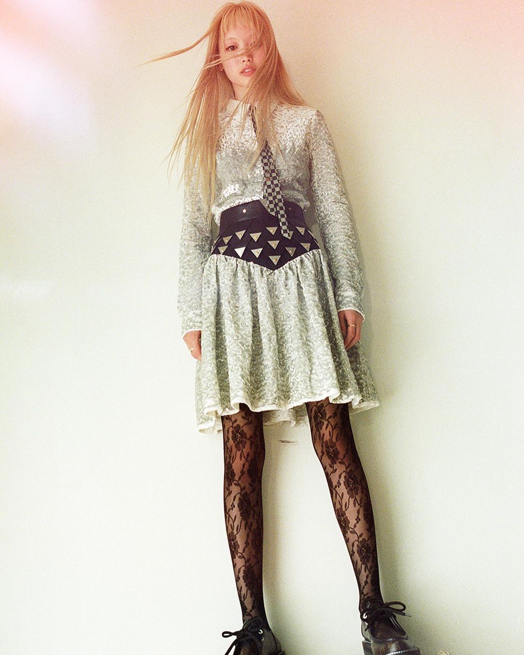 Young adult woman in long-sleeved silver dress with black tights posing against a white wall in a high angle, eye-level shot.