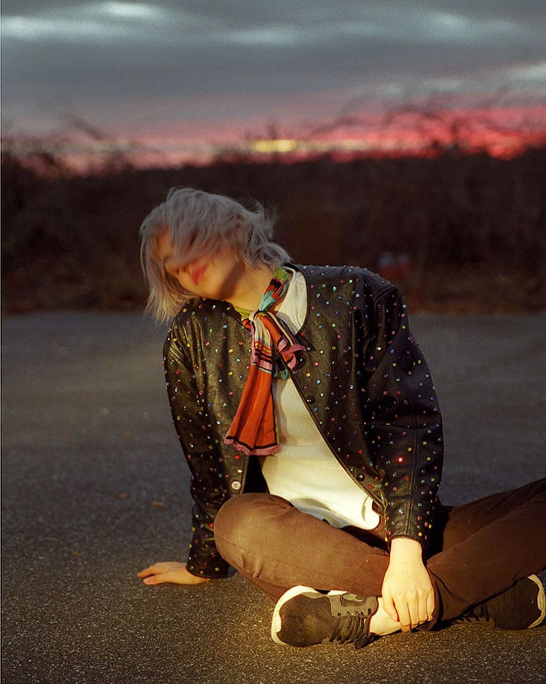 Woman in studded leather jacket sits cross-legged on asphalt. Sunset and bare trees in the background. Casual fashion shot.