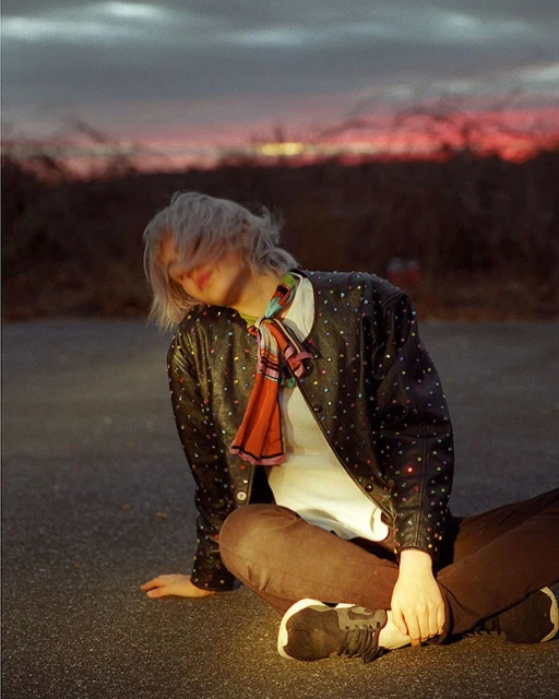 Woman in studded leather jacket sits cross-legged on asphalt. Sunset and bare trees in the background. Casual fashion shot.