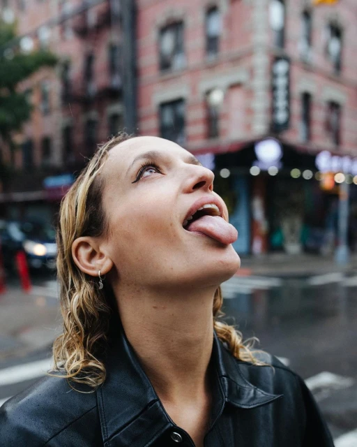 Adult woman outdoors in black leather jacket, standing on street, looking up with tongue out at urban building in daytime.