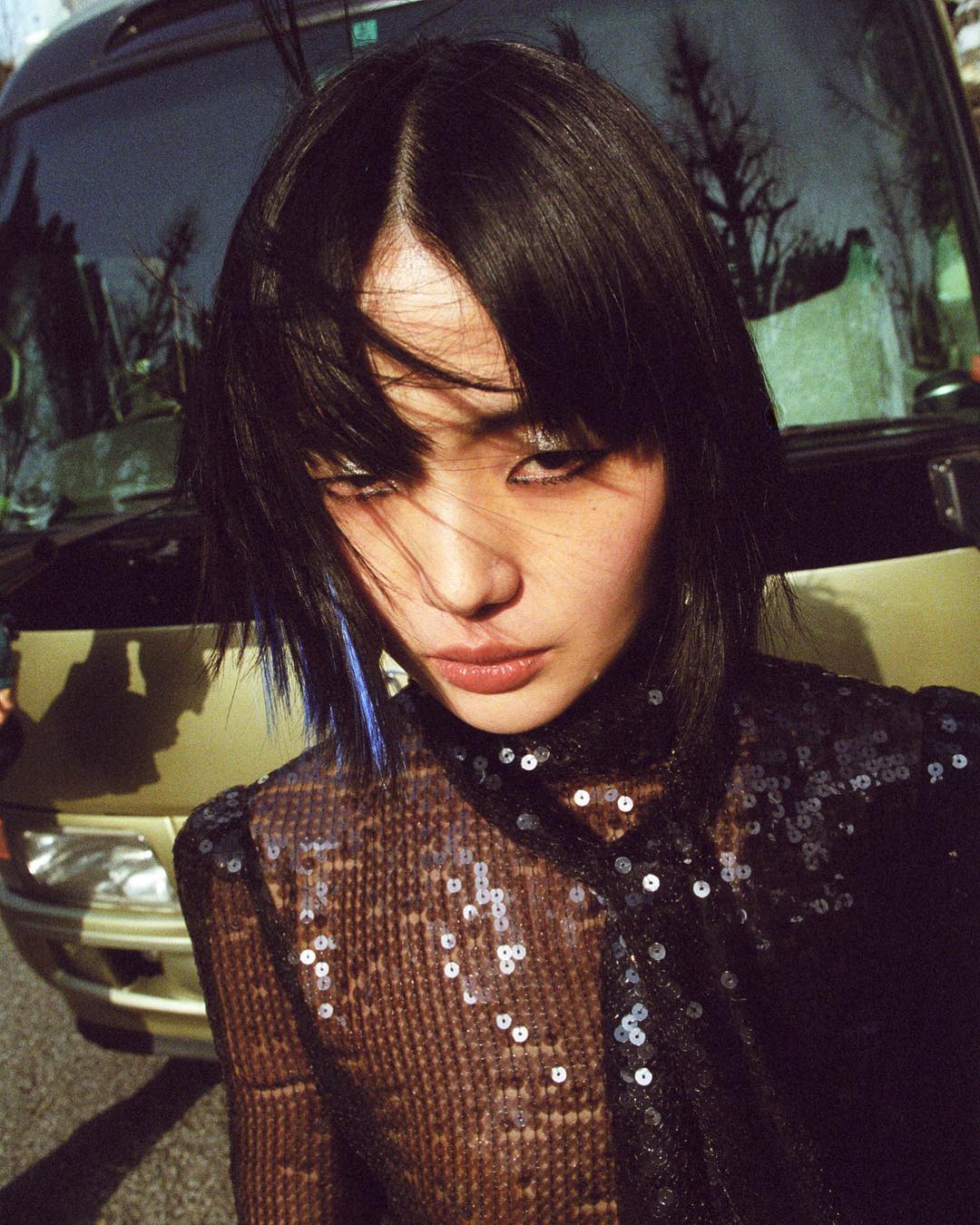 Portrait of an Asian woman with blue-streaked black hair wearing a sequined top outdoors with a bus in the background on a sunny day.