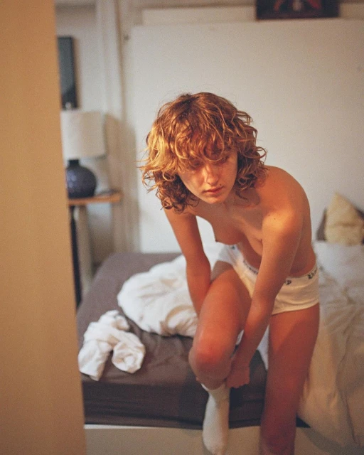 Close-up shot of a young adult female sitting on a bed, putting on socks. Natural lighting illuminates the bedroom interior.