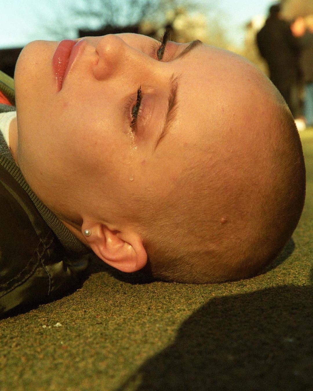 Close-up of a bald person with visible tear, lying down outside on green surface. Head and face are primary visual elements.