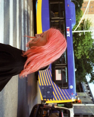 Woman with pink hair stands on a crosswalk next to a blue and yellow bus, trees and building in the background. Vertical orientation.
