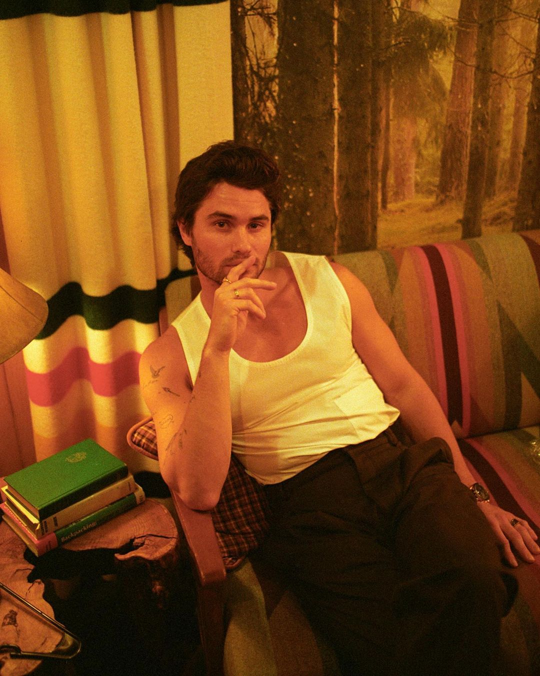 Adult male in a sleeveless shirt sitting indoors on a colorful couch with a forest background and books, showcasing casual style.