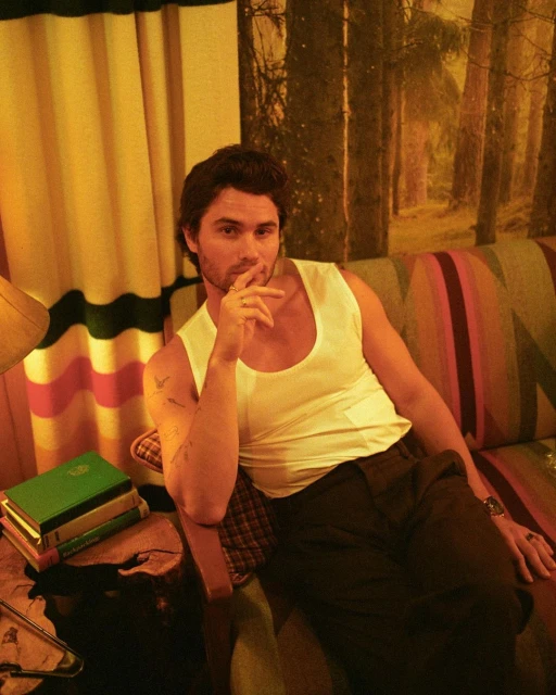 Adult male in a sleeveless shirt sitting indoors on a colorful couch with a forest background and books, showcasing casual style.