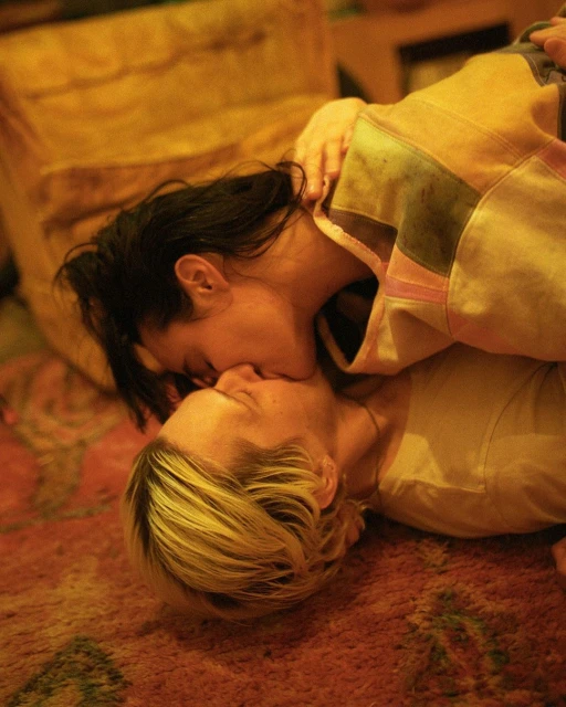 Close-up of two women kissing intimately on a patterned rug. The scene is lit with warm, golden-toned lighting.