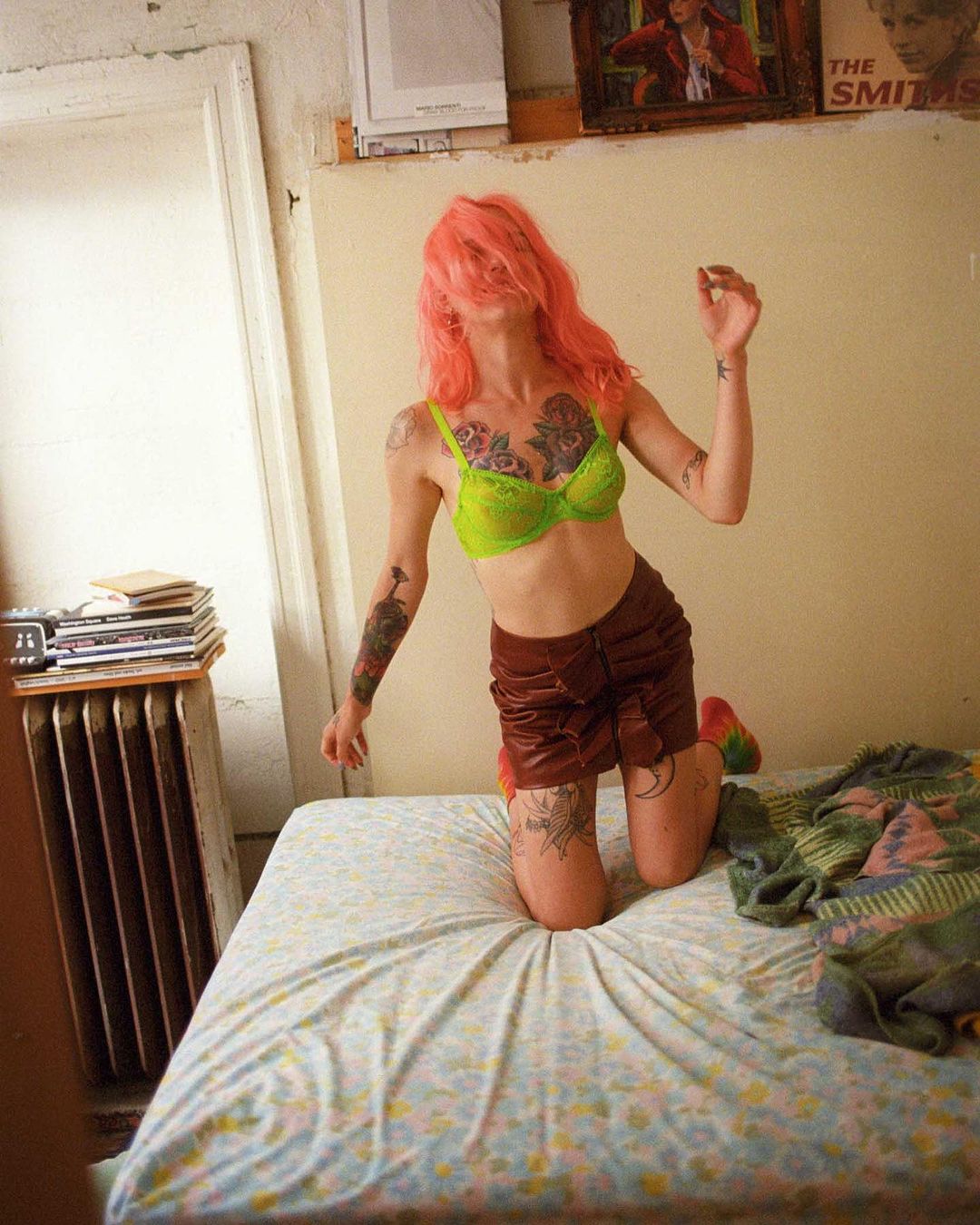 Woman in bra and skirt kneeling on bed with pink hair obscuring face, radiator and artwork in background of candid indoor shot.