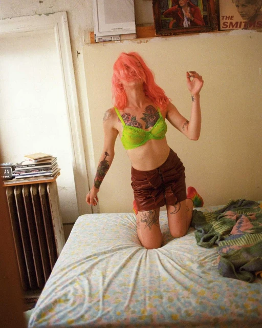 Woman in bra and skirt kneeling on bed with pink hair obscuring face, radiator and artwork in background of candid indoor shot.