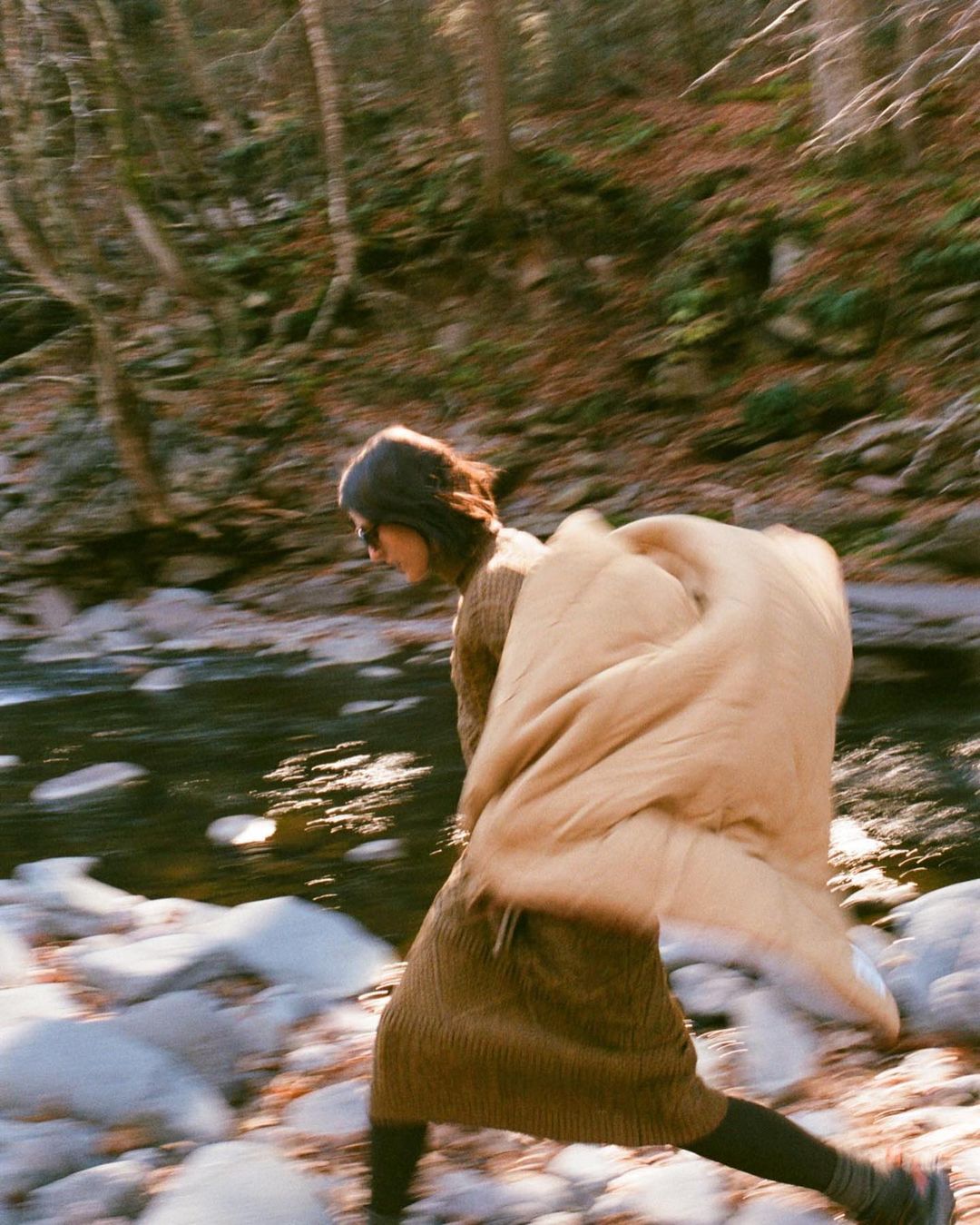 Woman crosses a shallow riverbed in a forest, carrying a tan puffer jacket or sleeping bag on her back in a dynamic, blurred motion.