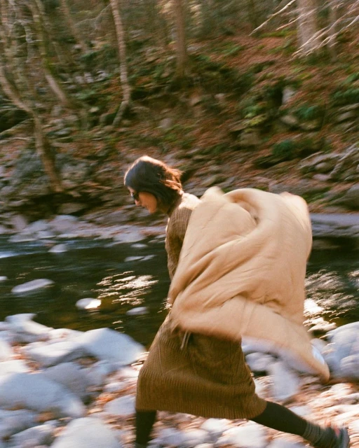 Woman crosses a shallow riverbed in a forest, carrying a tan puffer jacket or sleeping bag on her back in a dynamic, blurred motion.