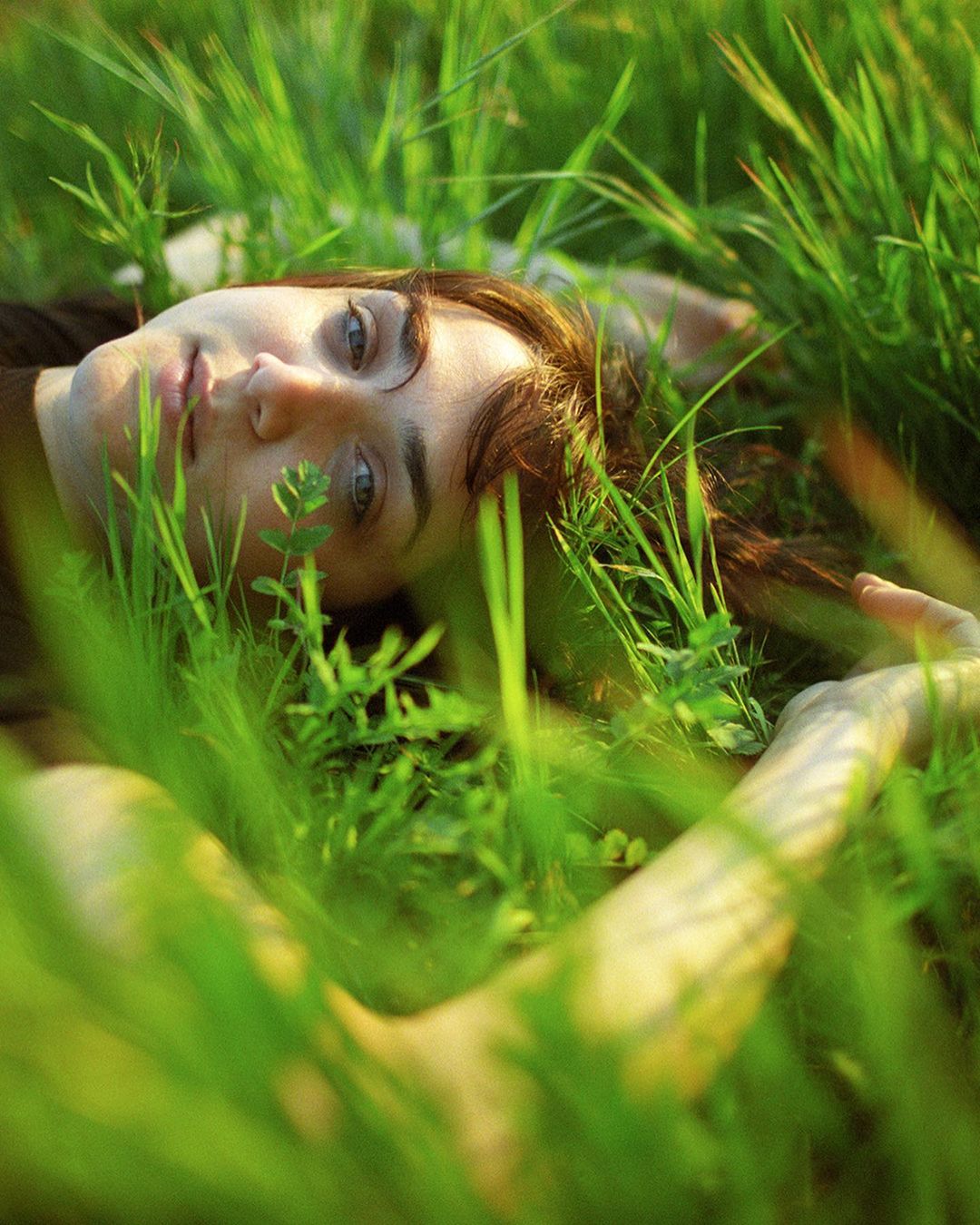 A woman with auburn hair lies in green grass, looking up at the camera with a serene expression in a natural outdoor setting.