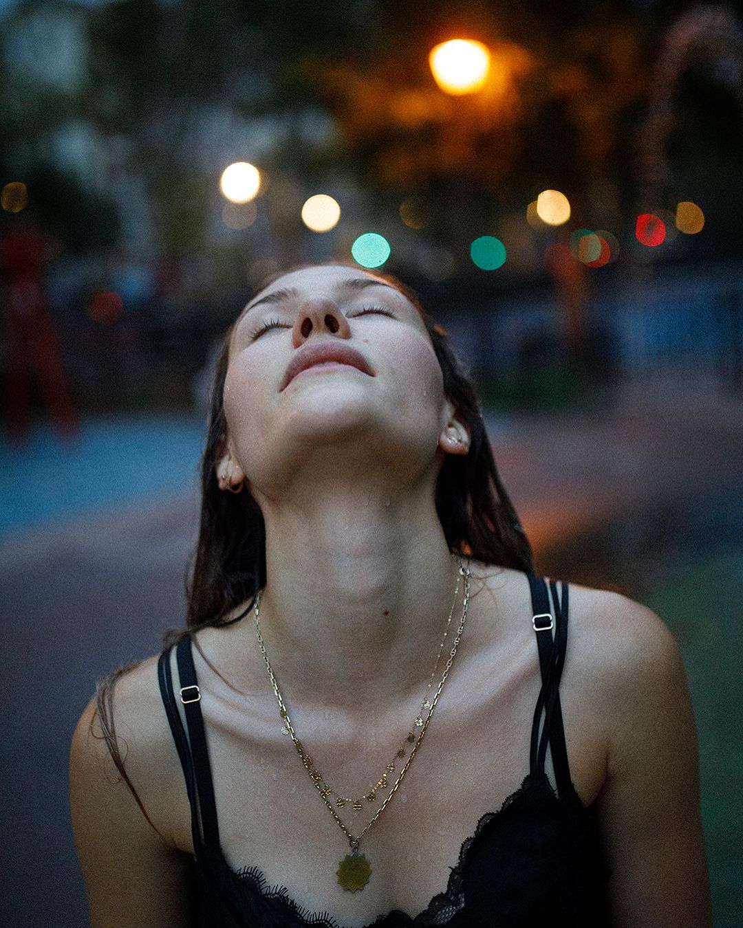 A woman looks up toward blurred city lights at night. She wears layered gold necklaces and a black lace tank top, with wet hair.