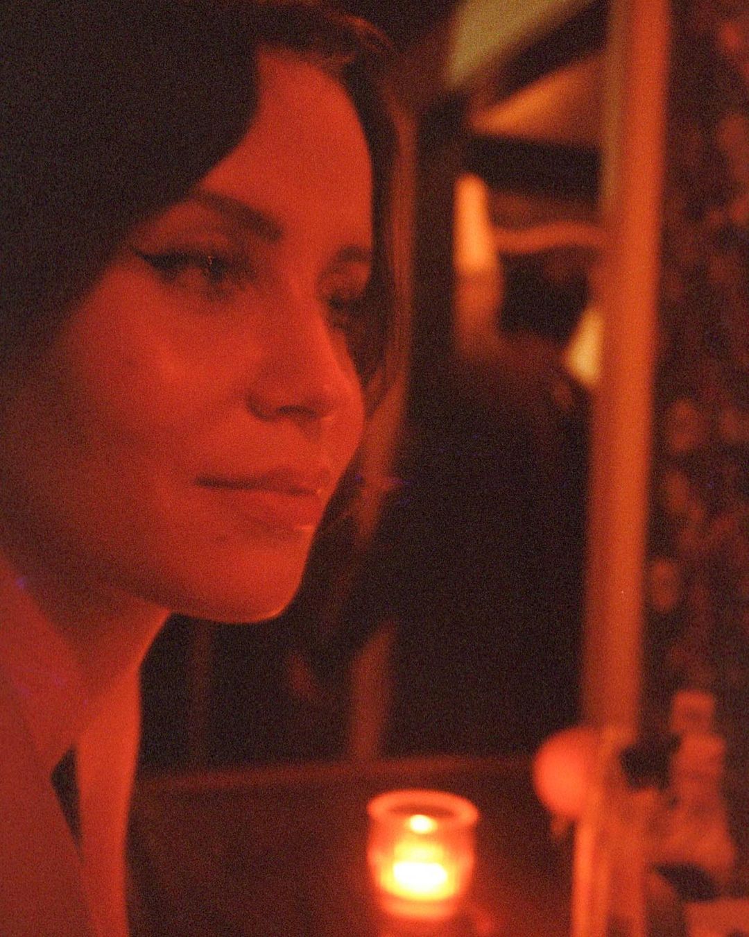 Portrait of a woman with dark hair and winged eyeliner, bathed in warm, orange-red light in a dimly lit indoor setting.