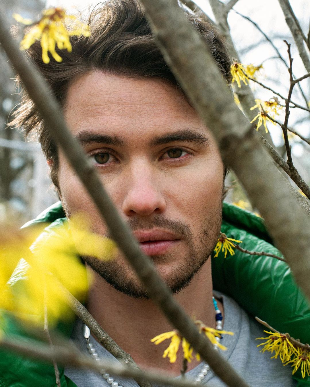 Close-up portrait of a man outdoors surrounded by flowering branches with a shallow depth of field focusing on his face and green jacket.
