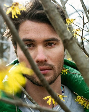 Close-up portrait of a man outdoors surrounded by flowering branches with a shallow depth of field focusing on his face and green jacket.