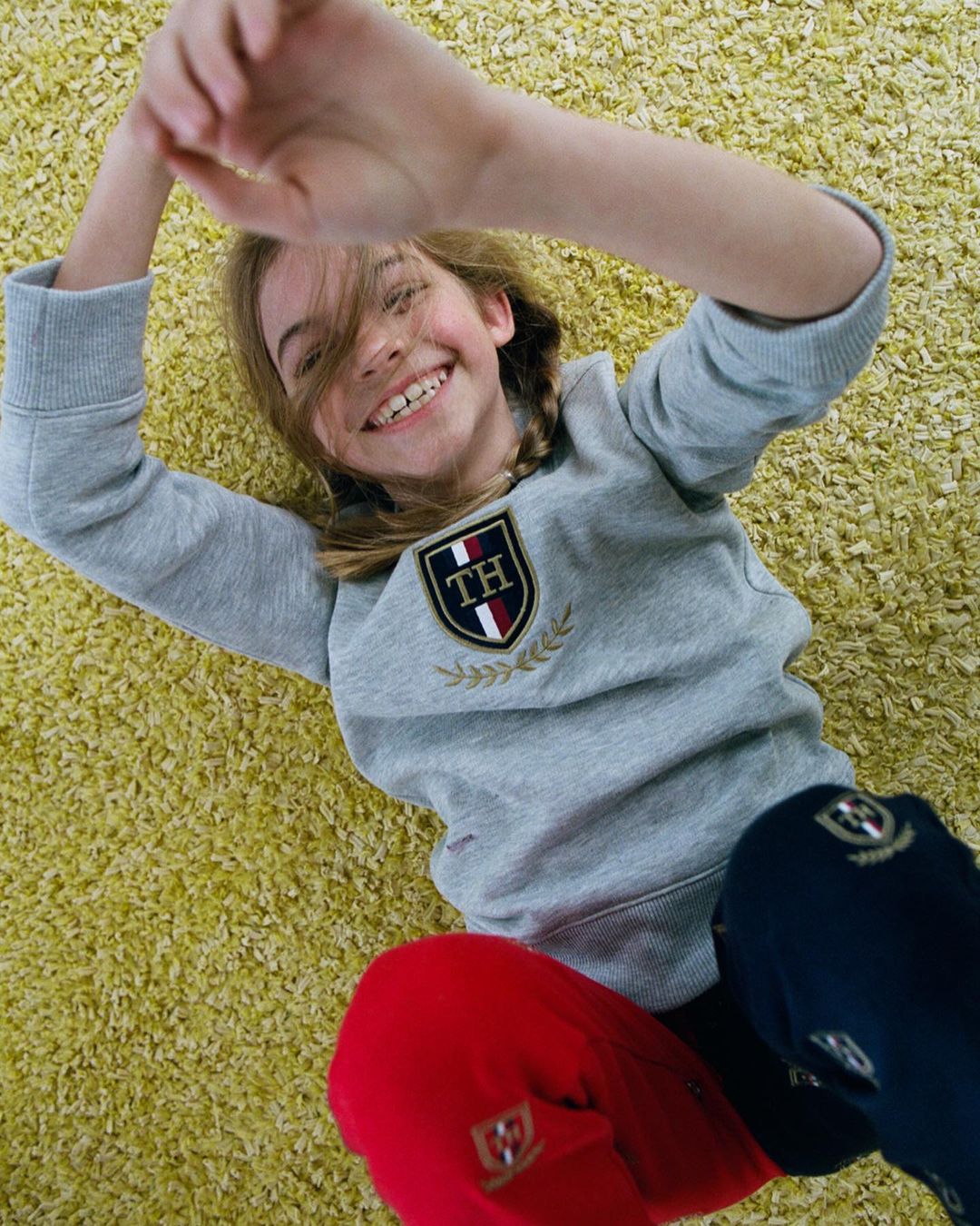 Smiling girl with braids, wearing a gray Tommy Hilfiger sweatshirt and red and blue pants, lies on a yellow shaggy carpet, looking up.