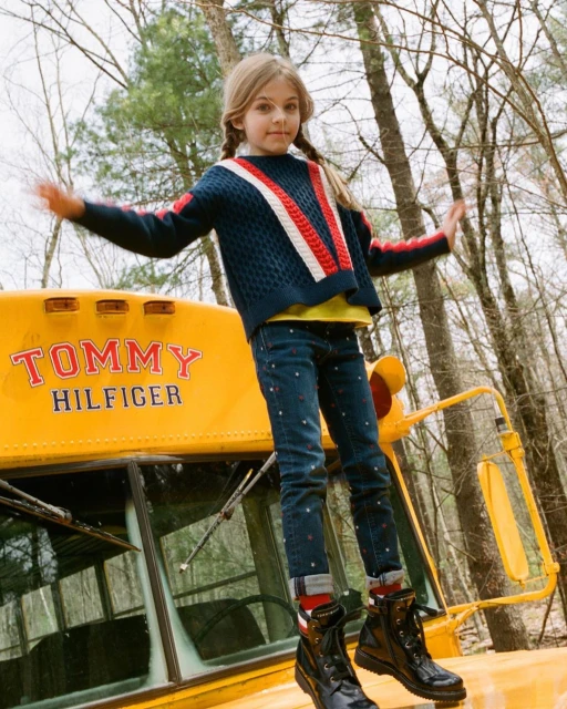 Young girl in Tommy Hilfiger sweater and jeans balancing on a yellow school bus in a wooded area, fashion campaign shoot.