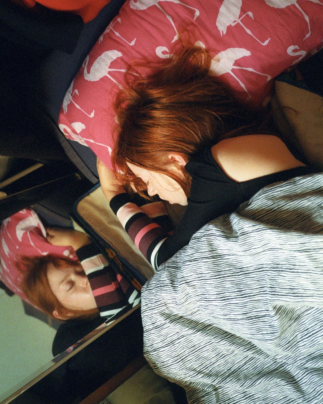 Red-haired woman is lying down on a bed with striped socks. A mirror reflects her face while she rests on a flamingo pillow.