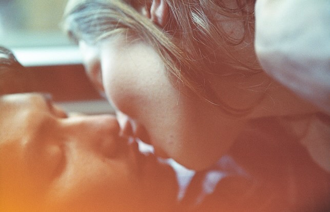 Close-up portrait of a couple with light skin tones kissing in soft, diffused lighting, captured with a shallow depth of field.