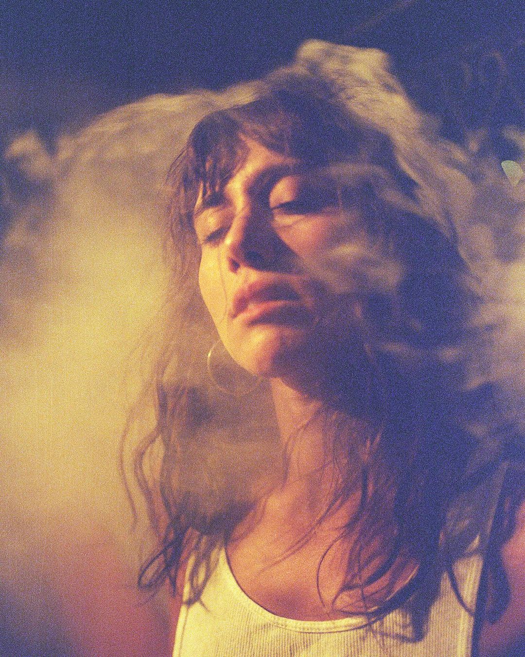 Portrait of a woman with long dark hair wearing a white tank top amidst swirling smoke and warm lighting.