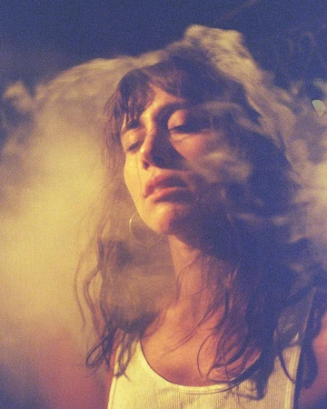 Portrait of a woman with long dark hair wearing a white tank top amidst swirling smoke and warm lighting.