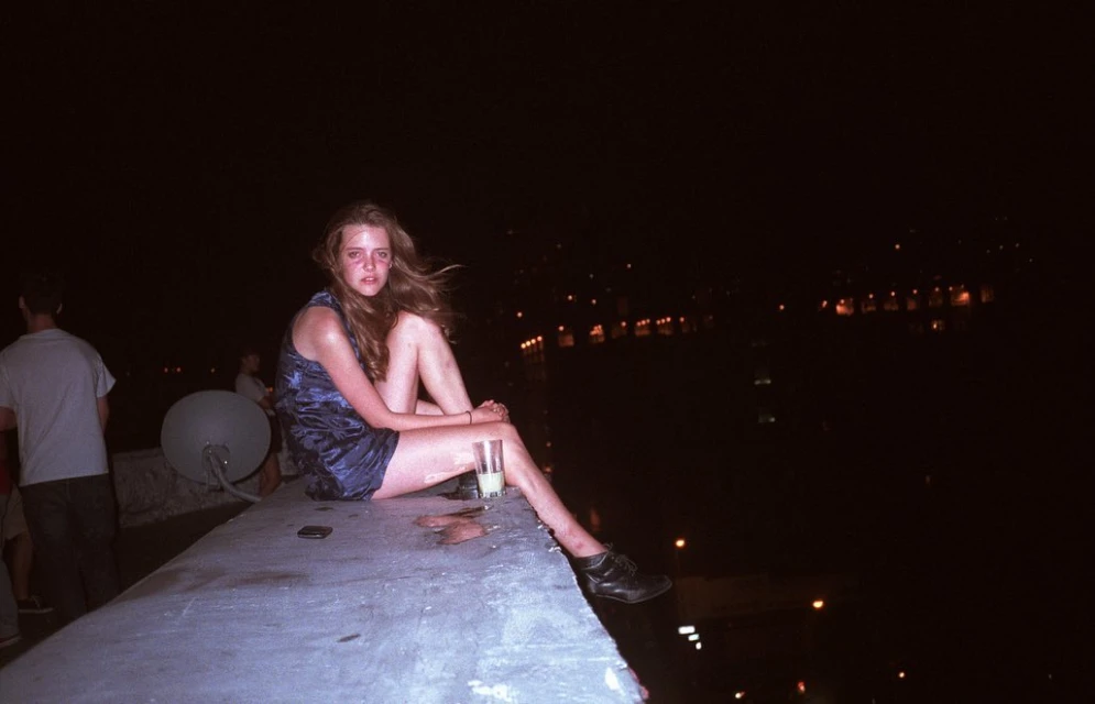 Woman in blue dress sits on concrete edge at night with city lights in background, holding a drink. Dark aesthetic, urban environment.