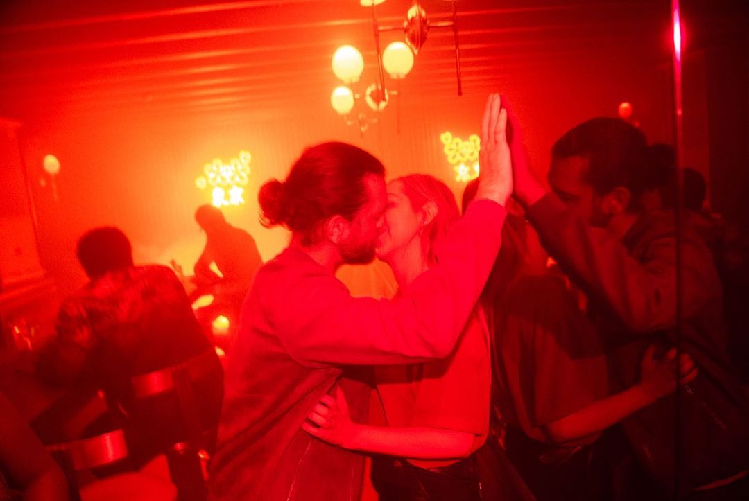 Couple kissing in a red-lit interior with other people; warm ambiance with diffused lighting, creating a vibrant and intimate atmosphere.