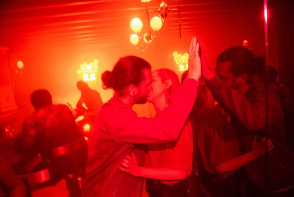 Couple kissing in a red-lit interior with other people; warm ambiance with diffused lighting, creating a vibrant and intimate atmosphere.