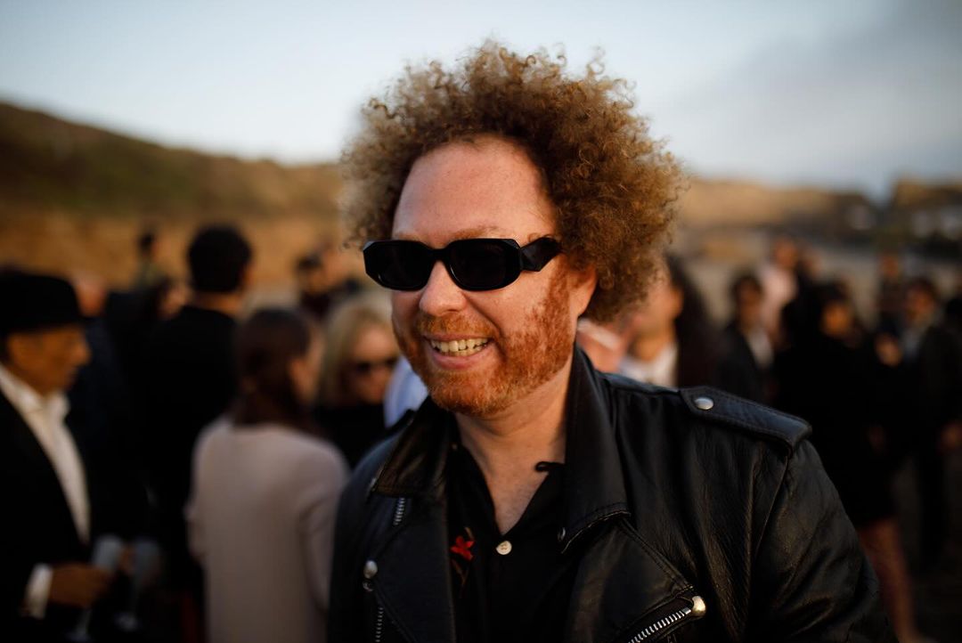 Smiling adult male with curly hair and sunglasses, wearing a black leather jacket in a soft outdoor light, surrounded by a crowd.