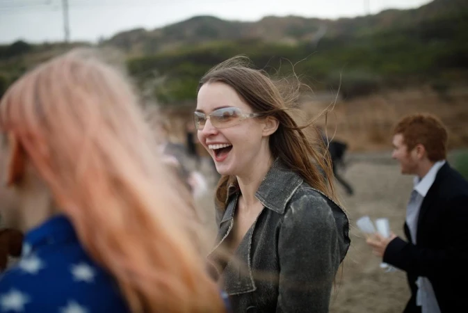 Laughing woman wearing sunglasses and a dark coat outdoors with other people in New York by Chad Moore