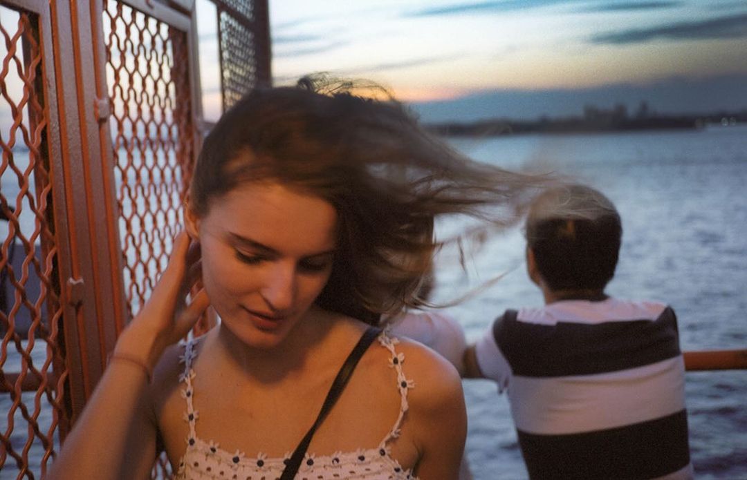 Young woman stands near the water at sunset, her hair blowing in the wind, with a striped-shirt figure in the background and metal fence.