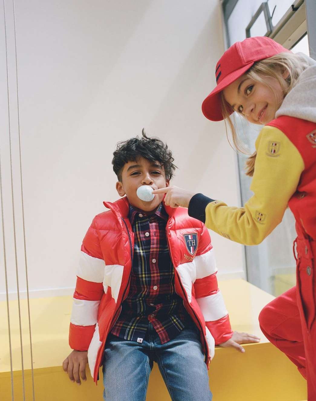 Two young children indoors. A boy in a red and white jacket has a white ball in his mouth. A girl in a red hat is visible behind him.