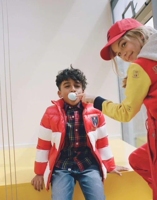 Two young children indoors. A boy in a red and white jacket has a white ball in his mouth. A girl in a red hat is visible behind him.