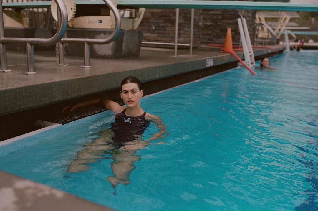 A woman in a swimsuit in a pool leans on the side with a diving board and ladder visible in the background. Pool water is blue.