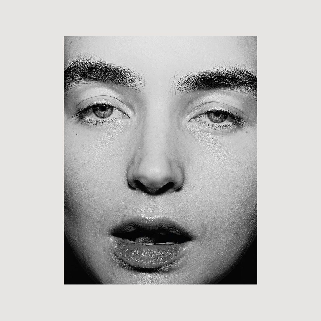 Close-up black-and-white portrait of an adult with open mouth, glossy lips, and neutral facial expression against a light neutral background.