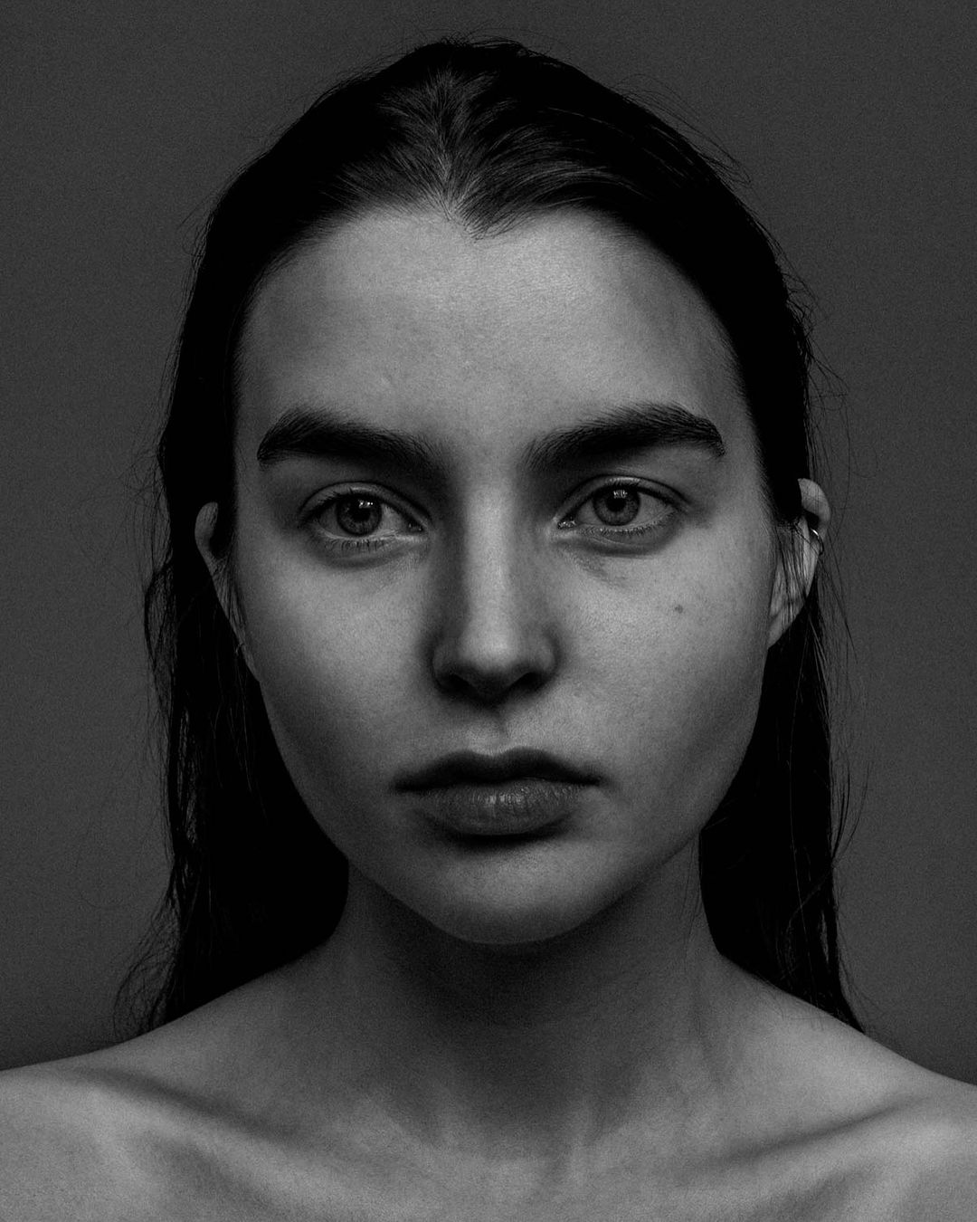 Monochrome close-up portrait of a young adult woman with dark hair, neutral facial expression, and simple jewelry against gray background.