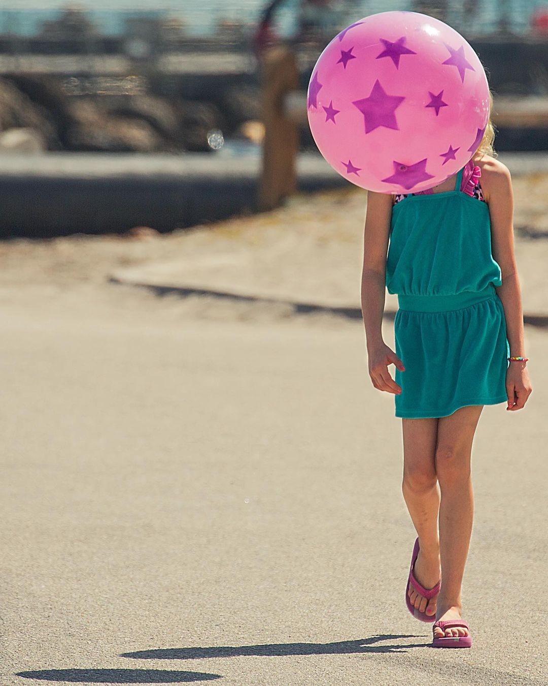 Child in a turquoise dress walks on a sidewalk with a pink beach ball with purple stars covering their face on a sunny day near a body of water.