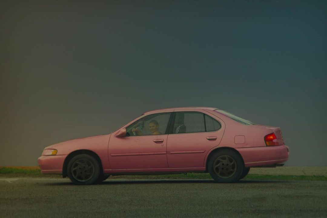 Side view of a pink sedan on an asphalt road, featuring an adult driver and a cloudy sky backdrop, creating a vintage travel scene.