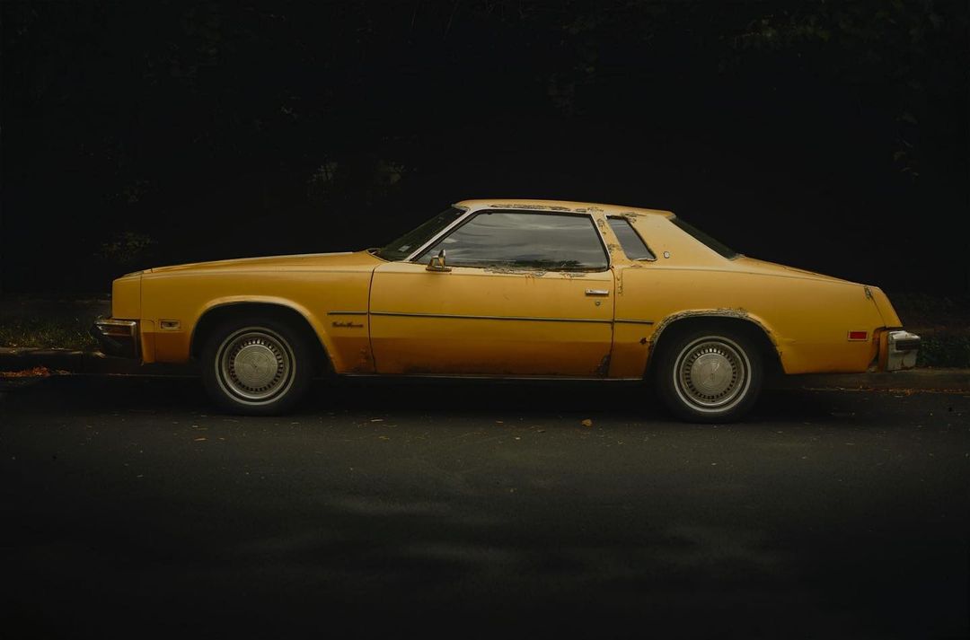 Profile view of a vintage yellow Cadillac coupe parked on the side of the road, set against a dark, leafy background.