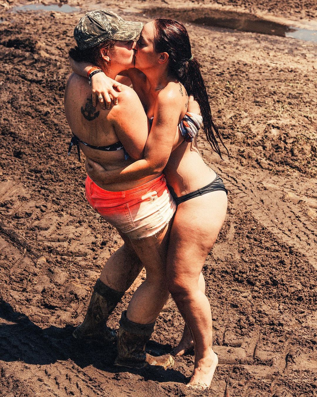 Two women in swimwear and boots embrace and kiss on a muddy surface on a sunny day, captured in a medium shot.